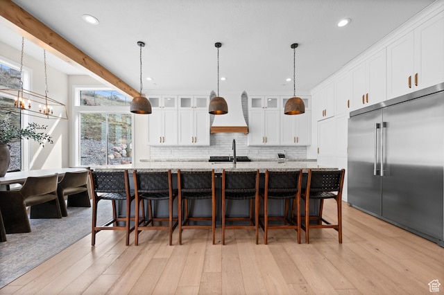 kitchen featuring custom exhaust hood, a large island with sink, light wood-style flooring, backsplash, and stainless steel built in refrigerator