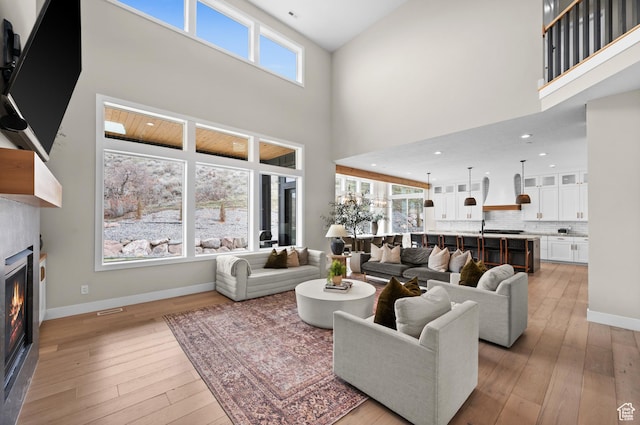 living area featuring a towering ceiling, a wealth of natural light, light wood finished floors, and a warm lit fireplace