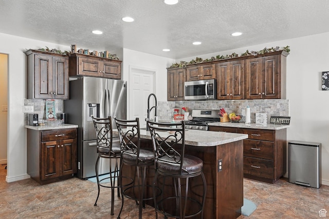 kitchen with tasteful backsplash, light countertops, appliances with stainless steel finishes, a kitchen island with sink, and dark brown cabinetry