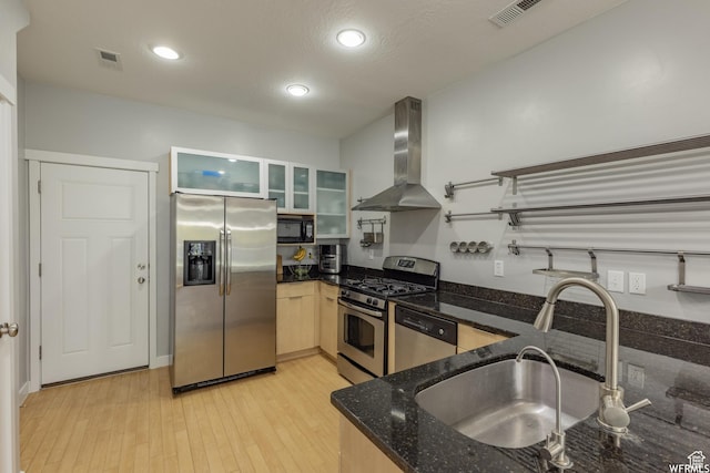kitchen with light wood-type flooring, dark stone countertops, wall chimney exhaust hood, stainless steel appliances, and a sink