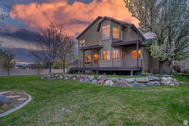 back of house at dusk with fence, a balcony, a yard, and stucco siding