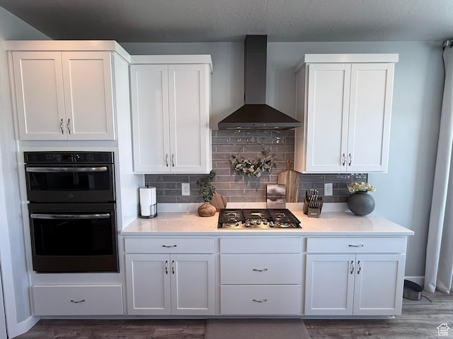 kitchen with dobule oven black, wall chimney range hood, stainless steel gas stovetop, white cabinets, and decorative backsplash
