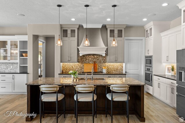 kitchen featuring light wood-type flooring, a center island with sink, premium range hood, backsplash, and appliances with stainless steel finishes