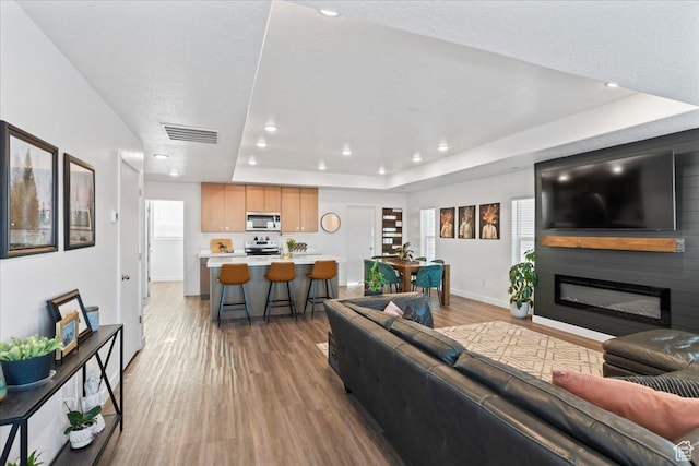 living room featuring light wood-style flooring, a glass covered fireplace, visible vents, recessed lighting, and a raised ceiling