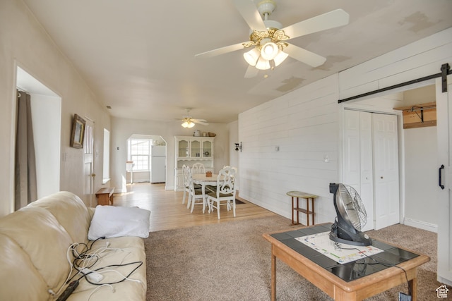 carpeted living area with arched walkways, wood walls, a ceiling fan, and a barn door