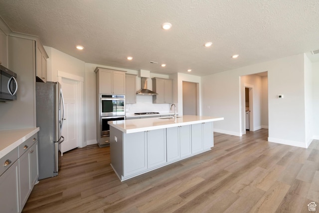 kitchen with wall chimney exhaust hood, a kitchen island with sink, a textured ceiling, gray cabinetry, and stainless steel appliances