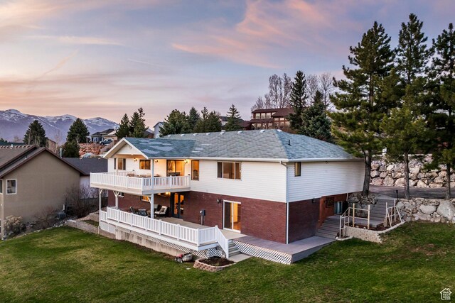 rear view of property with a lawn, roof with shingles, a deck with mountain view, brick siding, and a balcony