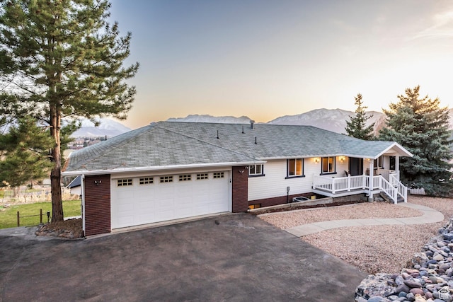 ranch-style home with a garage, covered porch, roof with shingles, a mountain view, and driveway