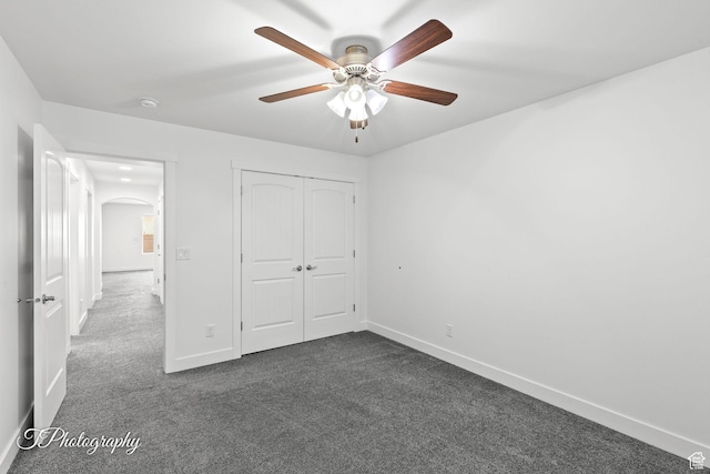 unfurnished bedroom with a closet, baseboards, arched walkways, dark carpet, and ceiling fan