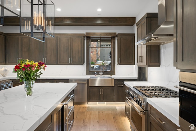 kitchen with a sink, wall chimney range hood, light wood-type flooring, appliances with stainless steel finishes, and dark brown cabinetry