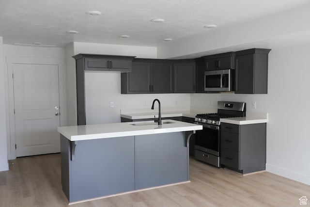 kitchen featuring a kitchen island with sink, a sink, stainless steel appliances, light wood finished floors, and baseboards