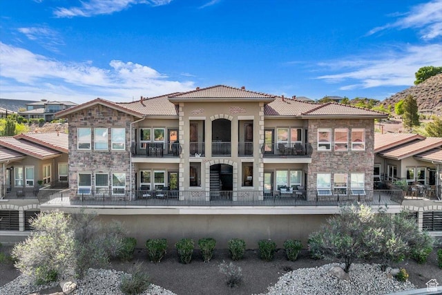 rear view of property featuring a tiled roof, stone siding, fence, stucco siding, and a balcony