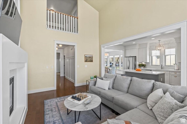 living room featuring baseboards, dark wood-style flooring, a chandelier, a towering ceiling, and a glass covered fireplace