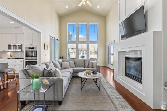 living area with a glass covered fireplace, a ceiling fan, a barn door, dark wood finished floors, and high vaulted ceiling