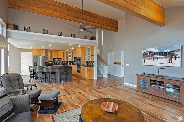 living room featuring a ceiling fan, stairs, a towering ceiling, beamed ceiling, and light wood finished floors