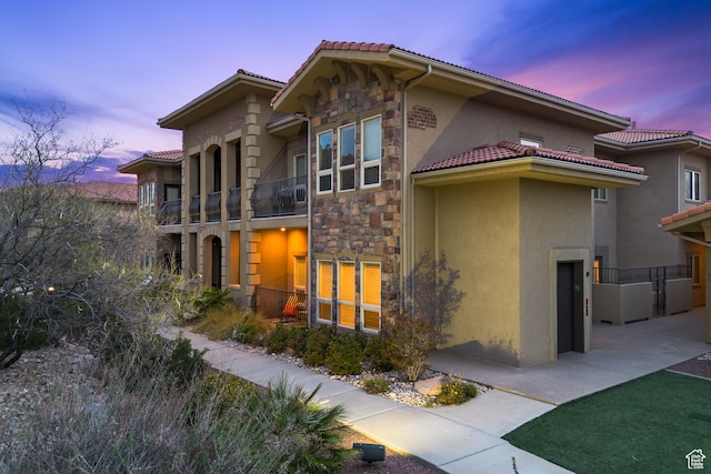 back of property at dusk featuring stone siding, a tiled roof, a balcony, and stucco siding