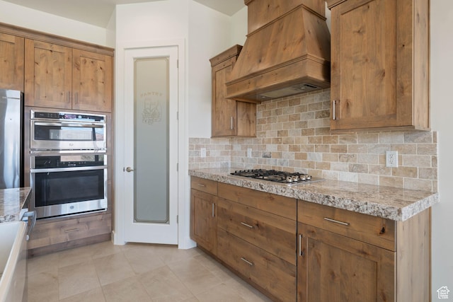 kitchen featuring stainless steel appliances, backsplash, light tile patterned floors, brown cabinetry, and custom exhaust hood
