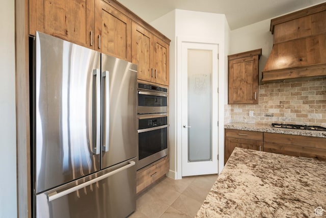 kitchen with light stone counters, brown cabinets, stainless steel appliances, custom range hood, and backsplash