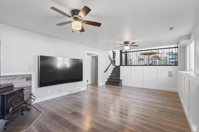 unfurnished living room featuring wood finished floors, stairs, ceiling fan, a wood stove, and visible vents