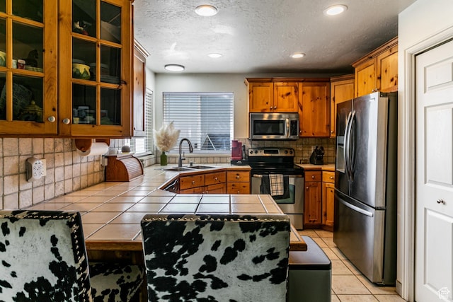 kitchen featuring a sink, tile countertops, brown cabinetry, light tile patterned floors, and appliances with stainless steel finishes