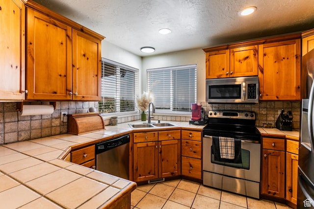 kitchen with tile counters, a sink, a textured ceiling, brown cabinets, and stainless steel appliances