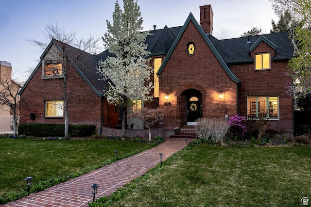english style home featuring a chimney, a front yard, roof with shingles, and brick siding