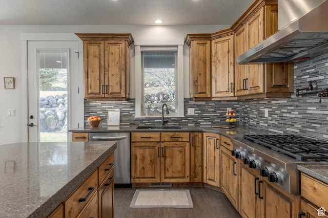 kitchen with appliances with stainless steel finishes, wall chimney range hood, a sink, brown cabinetry, and recessed lighting