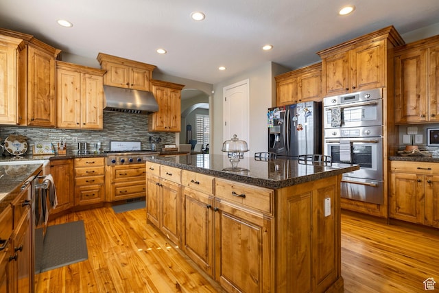 kitchen featuring under cabinet range hood, arched walkways, a warming drawer, a kitchen island, and appliances with stainless steel finishes
