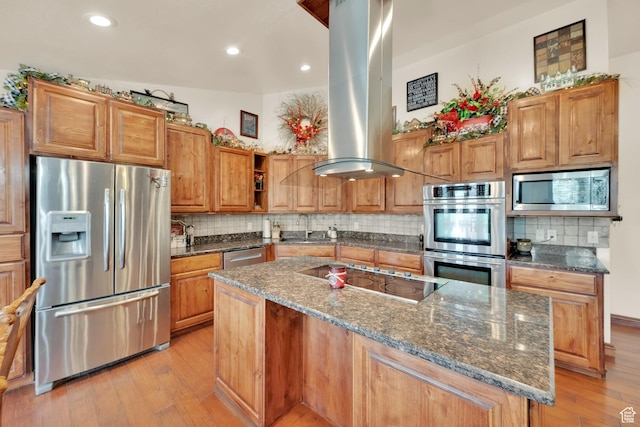 kitchen with dark stone countertops, island range hood, appliances with stainless steel finishes, light wood finished floors, and a center island with sink