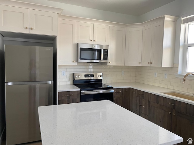 kitchen featuring stainless steel appliances, a sink, dark brown cabinets, backsplash, and light stone countertops