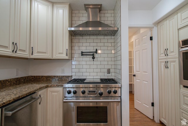 kitchen with stainless steel appliances, backsplash, wall chimney exhaust hood, dark stone counters, and light wood-style flooring