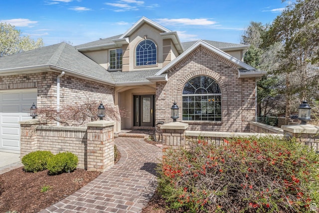 view of exterior entry with stucco siding, an attached garage, brick siding, and a shingled roof