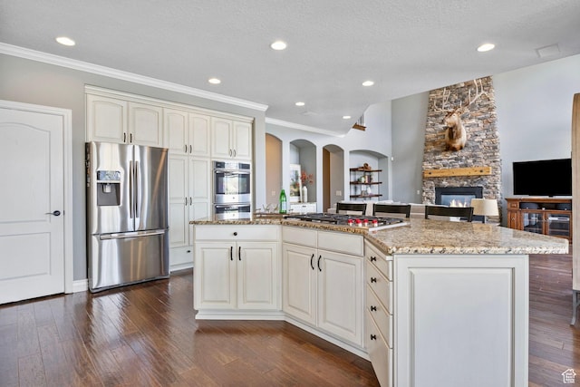 kitchen with stainless steel appliances, a stone fireplace, arched walkways, light stone countertops, and open floor plan
