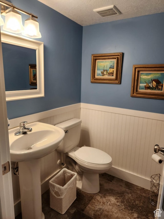 bathroom with a wainscoted wall, toilet, stone finish floors, and a textured ceiling