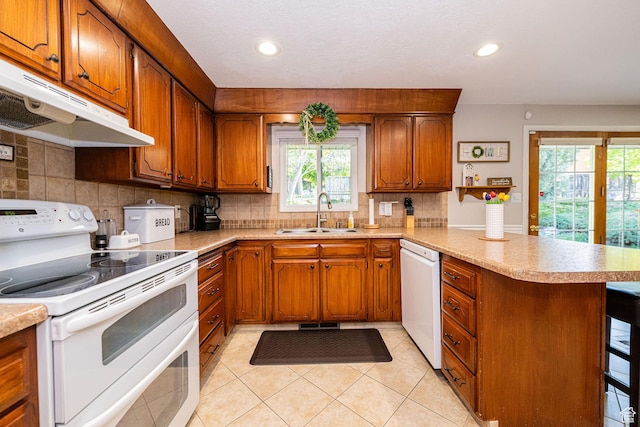 kitchen featuring a peninsula, white appliances, under cabinet range hood, brown cabinets, and a sink