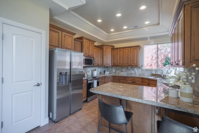 kitchen with appliances with stainless steel finishes, a tray ceiling, brown cabinetry, backsplash, and a peninsula