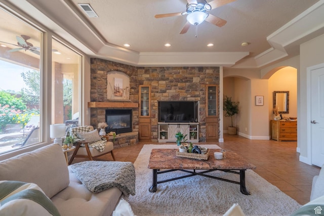 tiled living area with ceiling fan, arched walkways, a tray ceiling, a stone fireplace, and recessed lighting