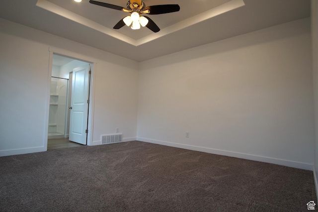 empty room with a tray ceiling, dark carpet, visible vents, a ceiling fan, and baseboards