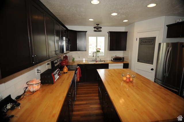 kitchen featuring a textured ceiling, backsplash, dark cabinetry, wooden counters, and appliances with stainless steel finishes