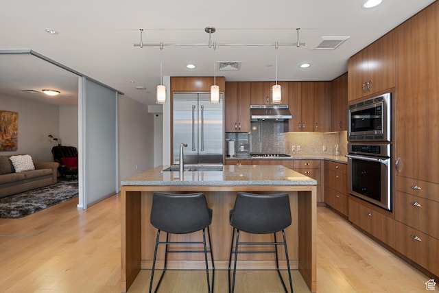 kitchen with brown cabinets, under cabinet range hood, visible vents, a sink, and built in appliances