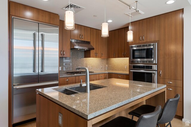 kitchen featuring under cabinet range hood, visible vents, a sink, brown cabinetry, and built in appliances