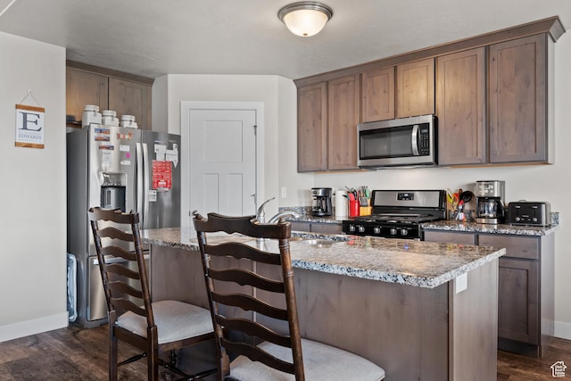 kitchen with an island with sink, baseboards, dark wood-type flooring, and appliances with stainless steel finishes