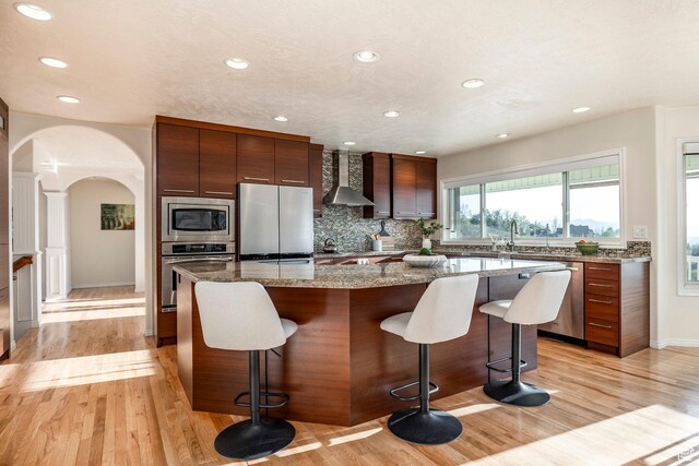 kitchen with stainless steel appliances, a breakfast bar, arched walkways, wall chimney exhaust hood, and a center island