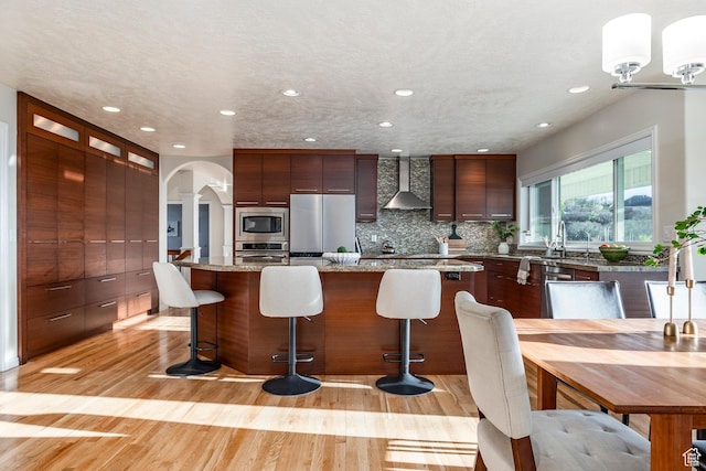 kitchen featuring recessed lighting, backsplash, wall chimney range hood, arched walkways, and appliances with stainless steel finishes