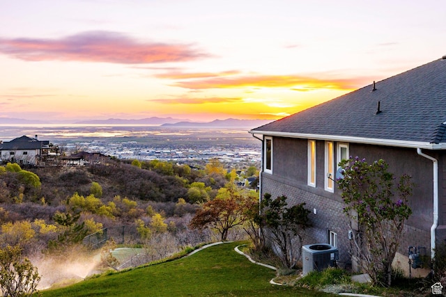 property exterior at dusk with a mountain view, central AC, brick siding, a shingled roof, and a yard