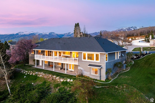 back of house at dusk featuring a yard, a patio area, roof with shingles, and a deck with mountain view