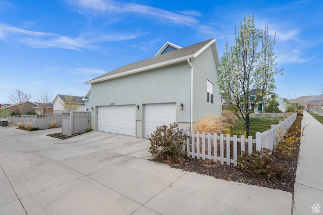 view of side of home with driveway, an attached garage, fence, stucco siding, and roof with shingles