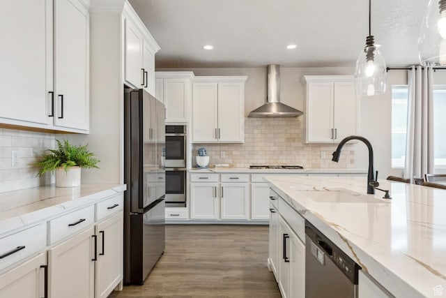 kitchen with stainless steel appliances, a sink, wall chimney exhaust hood, pendant lighting, and white cabinetry