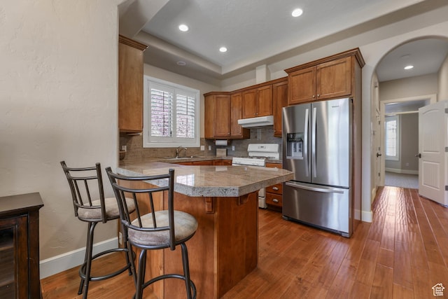 kitchen featuring white gas stove, a sink, brown cabinetry, stainless steel refrigerator with ice dispenser, and under cabinet range hood