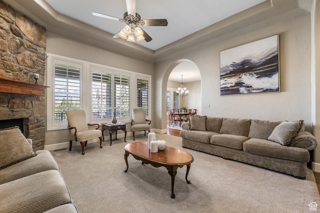 carpeted living area with arched walkways, a stone fireplace, baseboards, and ceiling fan with notable chandelier